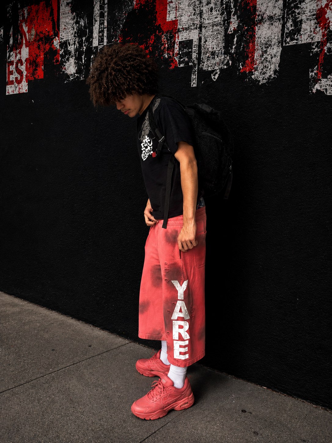 Young man with an afro wearing the salmon RELIC 3/4 shorts by YARE Clothing against a red and black street poster wall